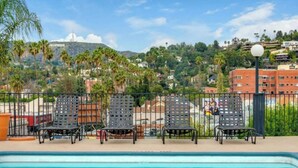 A poolside area with four metal chairs and a lamp post, overlooking a cityscape with a prominent hill and palm trees.