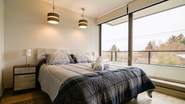 A modern bedroom with a large bed, two bedside lamps, a nightstand, and a view of trees through large windows. - Nuevo y hermoso depto central en Pucón (Pucon)