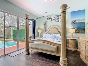 Ein Bett mit Baldachin, eine Blumendecke, ein Nachttisch mit Lampe, eine Kommode mit Spiegel und ein großes Fenster mit Blick auf einen Pool und Grünflächen.