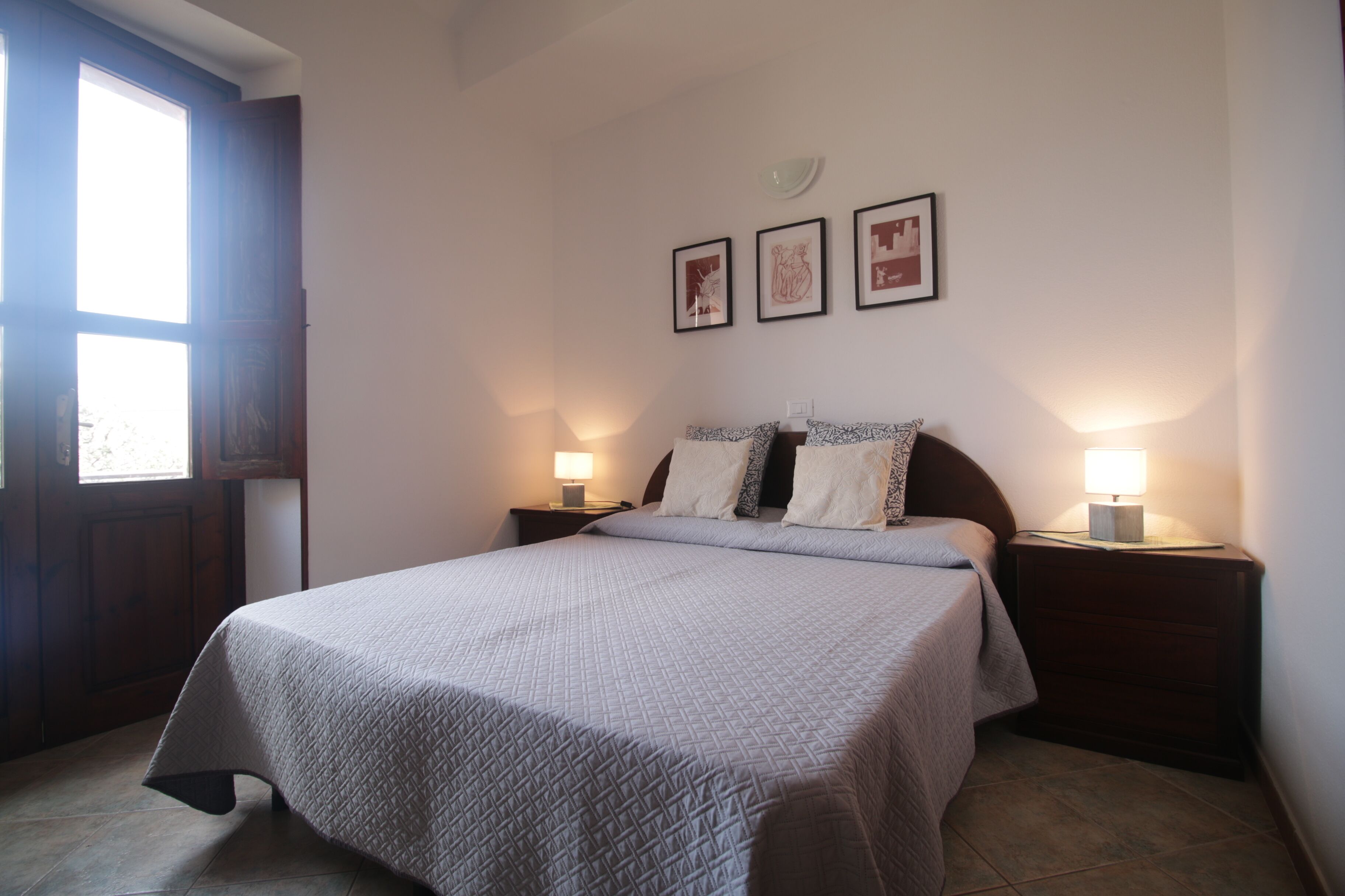 A bedroom with a bed, two bedside tables with lamps, a window, and three framed pictures on the wall.