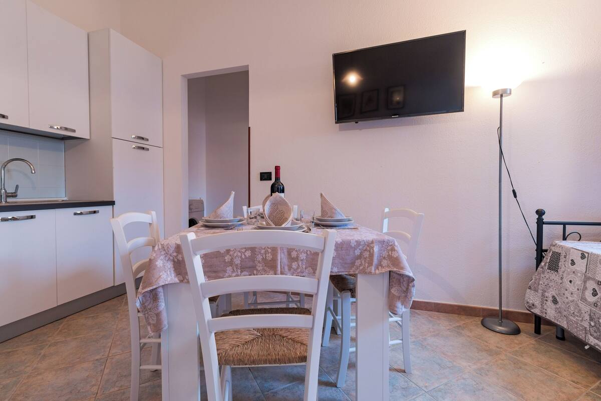 A dining area with a table set for four, a television mounted on the wall, and a kitchen area with cabinets and a countertop.