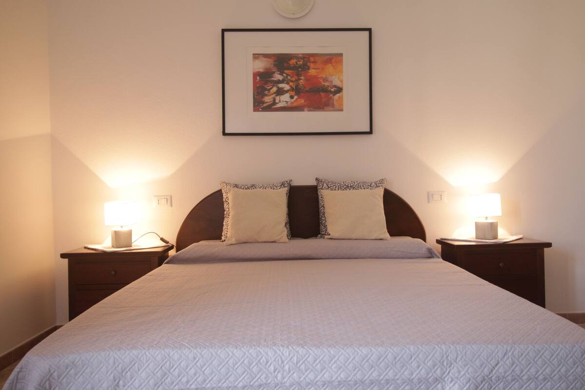A neatly made bed with a quilted cover, two bedside lamps, and a framed abstract painting on the wall.