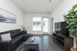 A modern living room with a black sectional sofa, a coffee table, a flat-screen TV, and a large window with blinds. - 2BR Hollywood Apt w/ Rooftop deck & Stunning Views (Los Angeles)