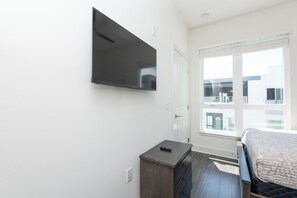 Une chambre à coucher avec un lit, un téléviseur fixé au mur, un petit bureau et une fenêtre donnant sur des immeubles.