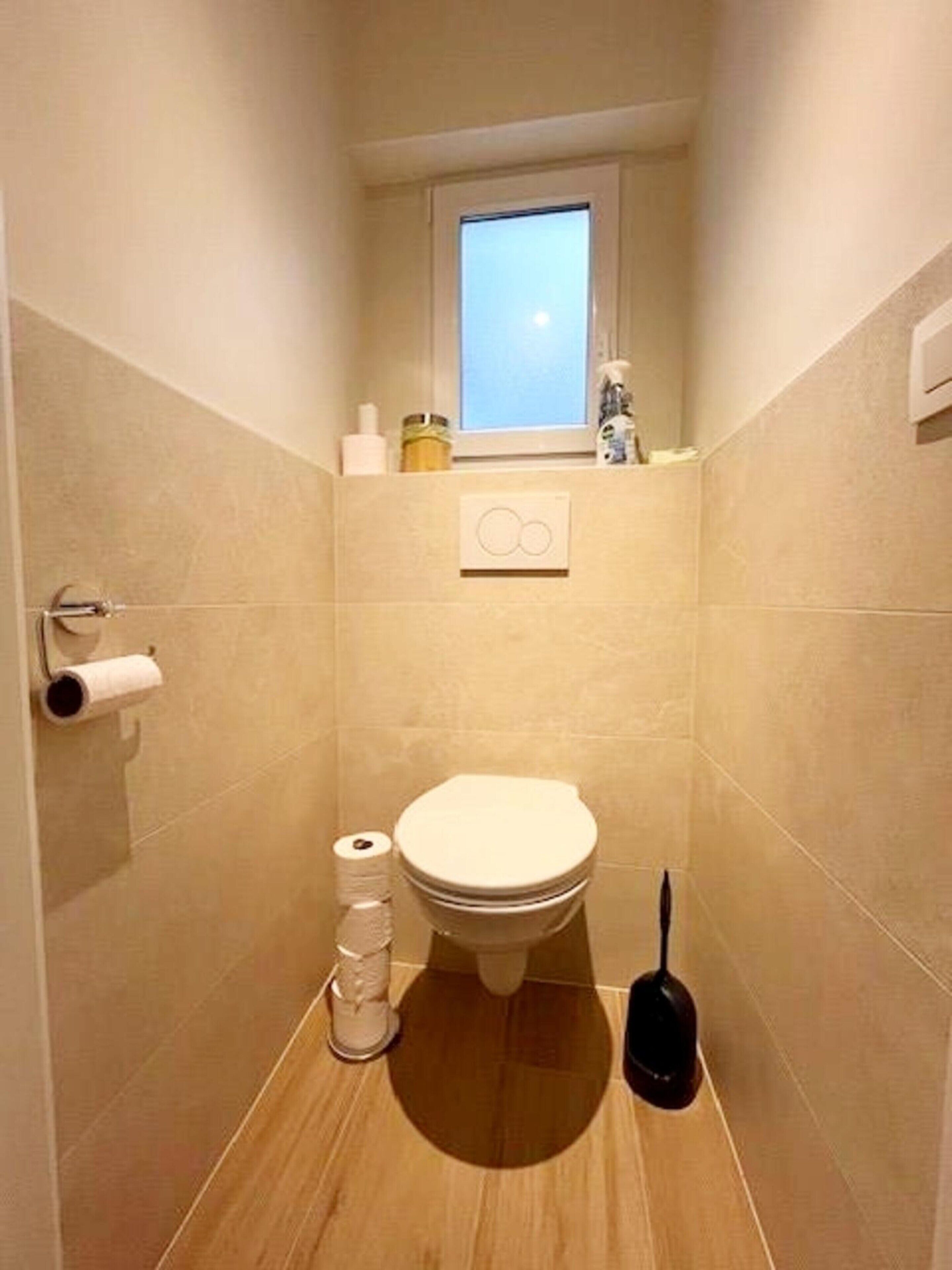 Ein kleines, schmales Badezimmer mit Toilette, einem Fenster und einem Ablagefach für Toilettenartikel.