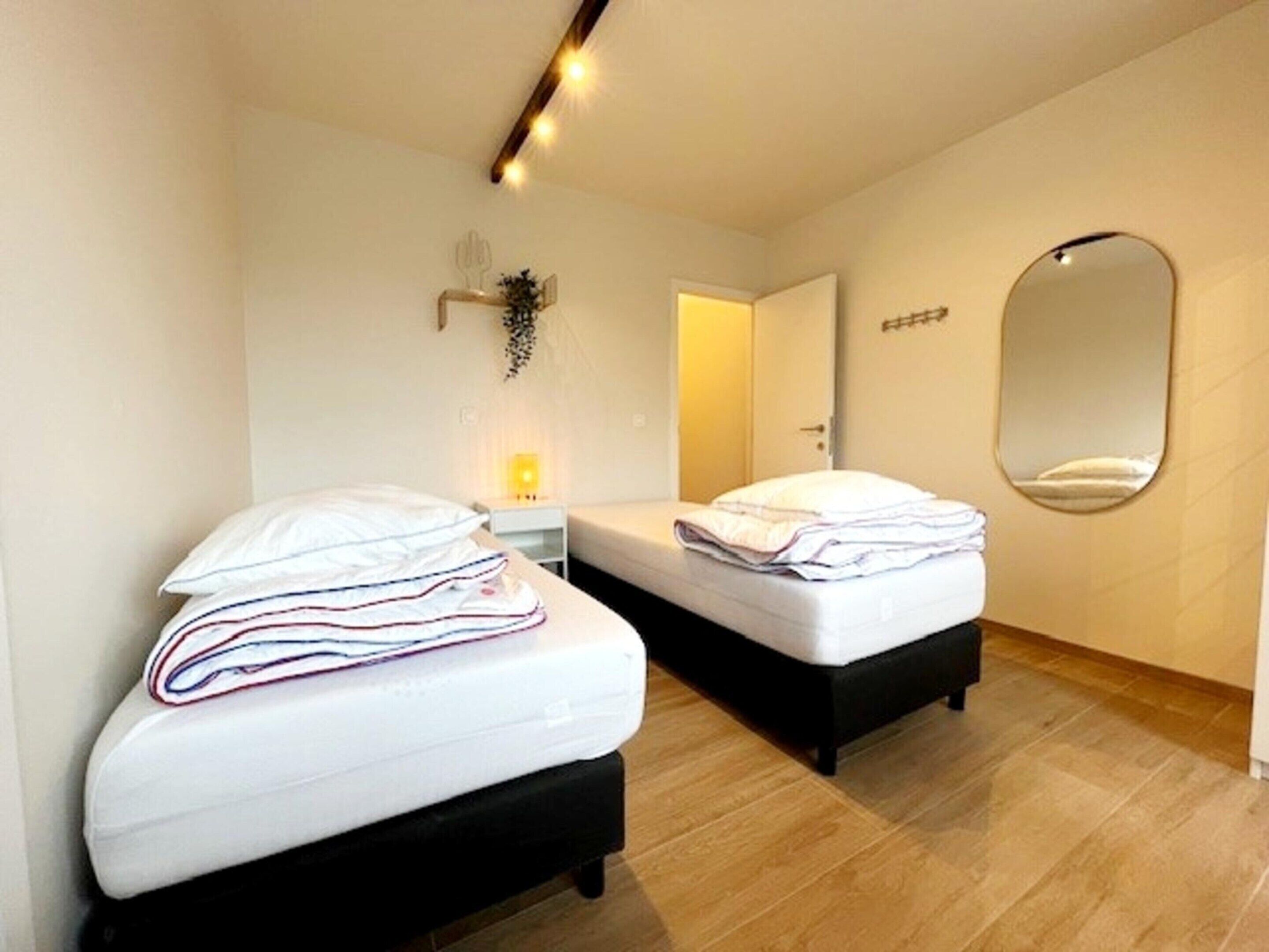 Ein Zimmer mit zwei Betten, einem Nachttisch mit Lampe, einem Spiegel an der Wand und einer Tür zu einem anderen Raum.