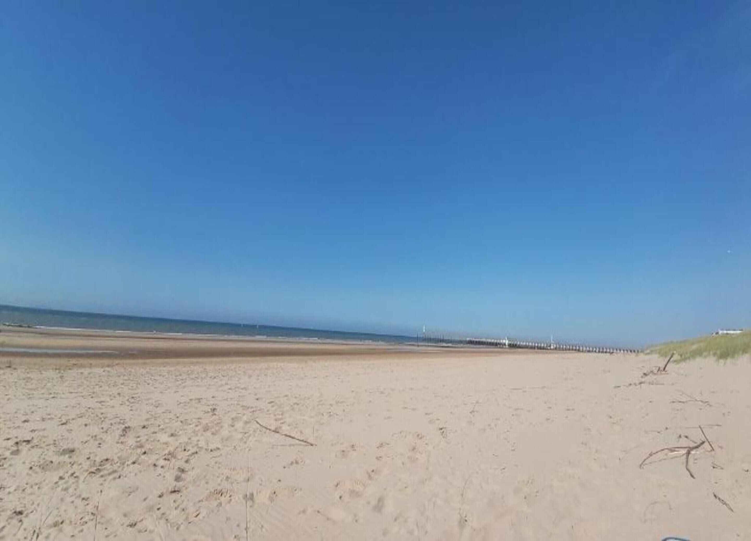 Une vaste plage de sable fin, un ciel bleu dégagé et une longue jetée qui s’avance dans l’eau.