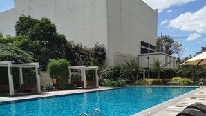 Una piscina con solárium empedrado, rodeada de vegetación y un edificio blanco.