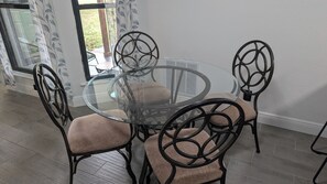 A round glass table with four chairs, each with a unique metal frame design, in a room with a tiled floor and a window with patterned curtains.