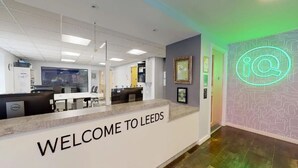 Ett välkomstområde med en skylt som säger ”VÄLKOMMEN TILL LEEDS”, en receptionsdisk och en neonbelyst skylt med texten ”i@” på väggen.