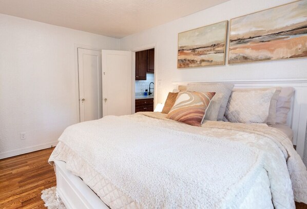 A bedroom with a large bed, a door to a bathroom, and a kitchen in the background.