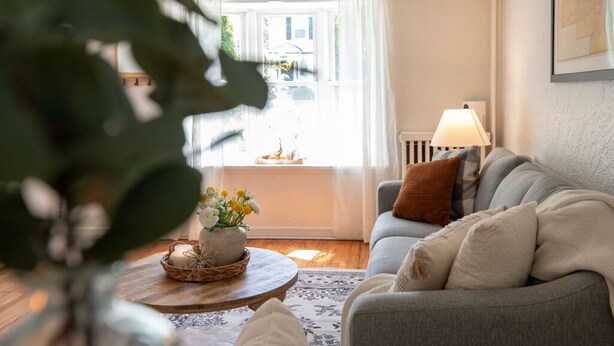 Un salon chaleureux avec un canapé gris, une table basse ronde en bois et une fenêtre ornée de rideaux blancs.