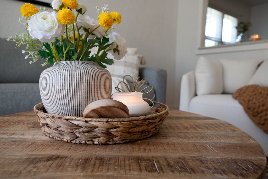 Een rieten mand met een bloemenvaas en een kaars op een houten tafel in een woonkamer.