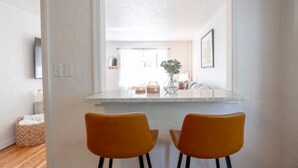 Une cuisine moderne avec un plan de travail blanc, deux chaises orange et une vue sur la salle à manger.