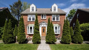 Une maison à deux étages, construite en briques, avec une entrée centrale, des fenêtres disposées de manière symétrique et une pelouse devant, agrémentée de haies taillées.