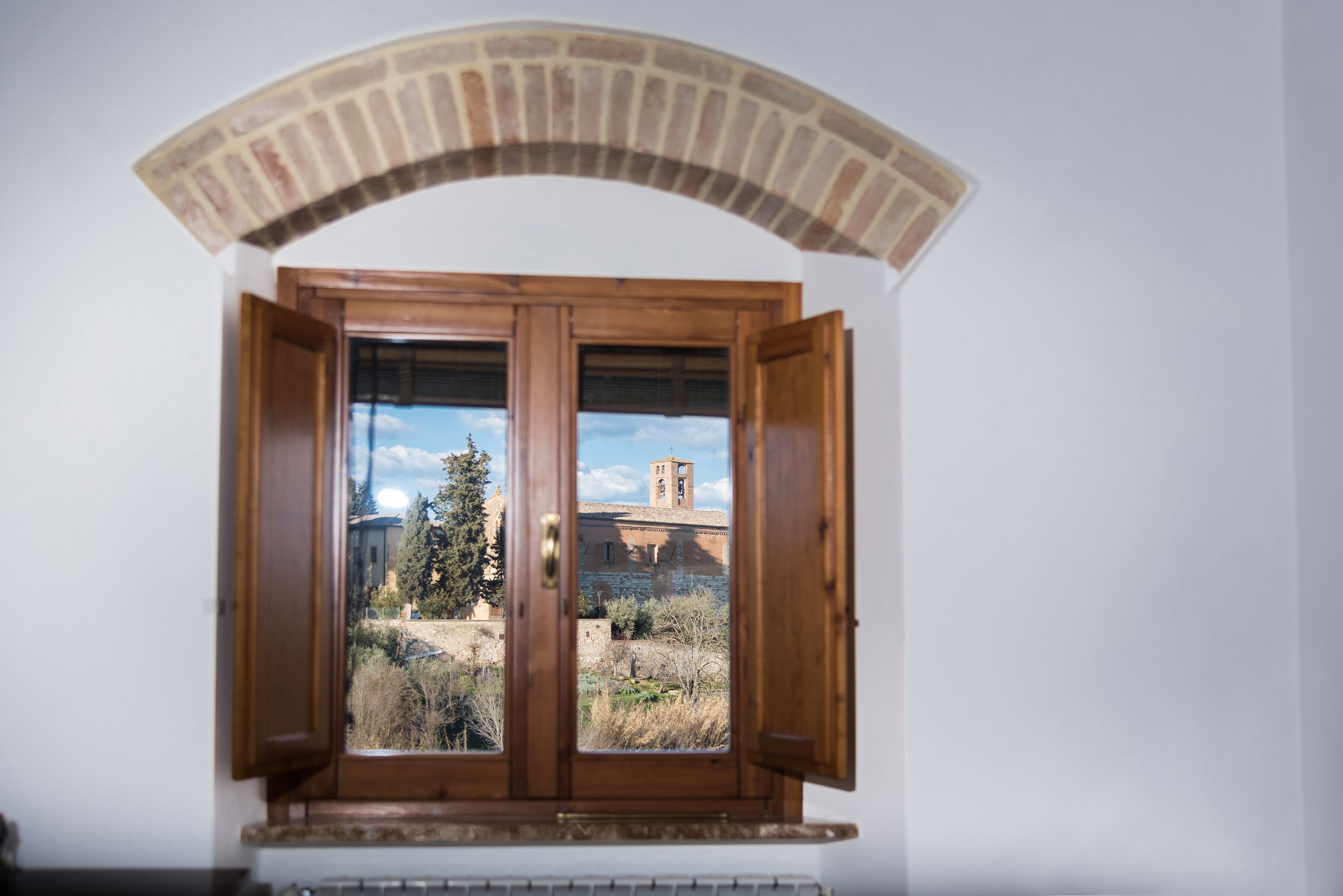 A room with a wooden window featuring a brick arch above it, providing a view of a building with a bell tower and trees.