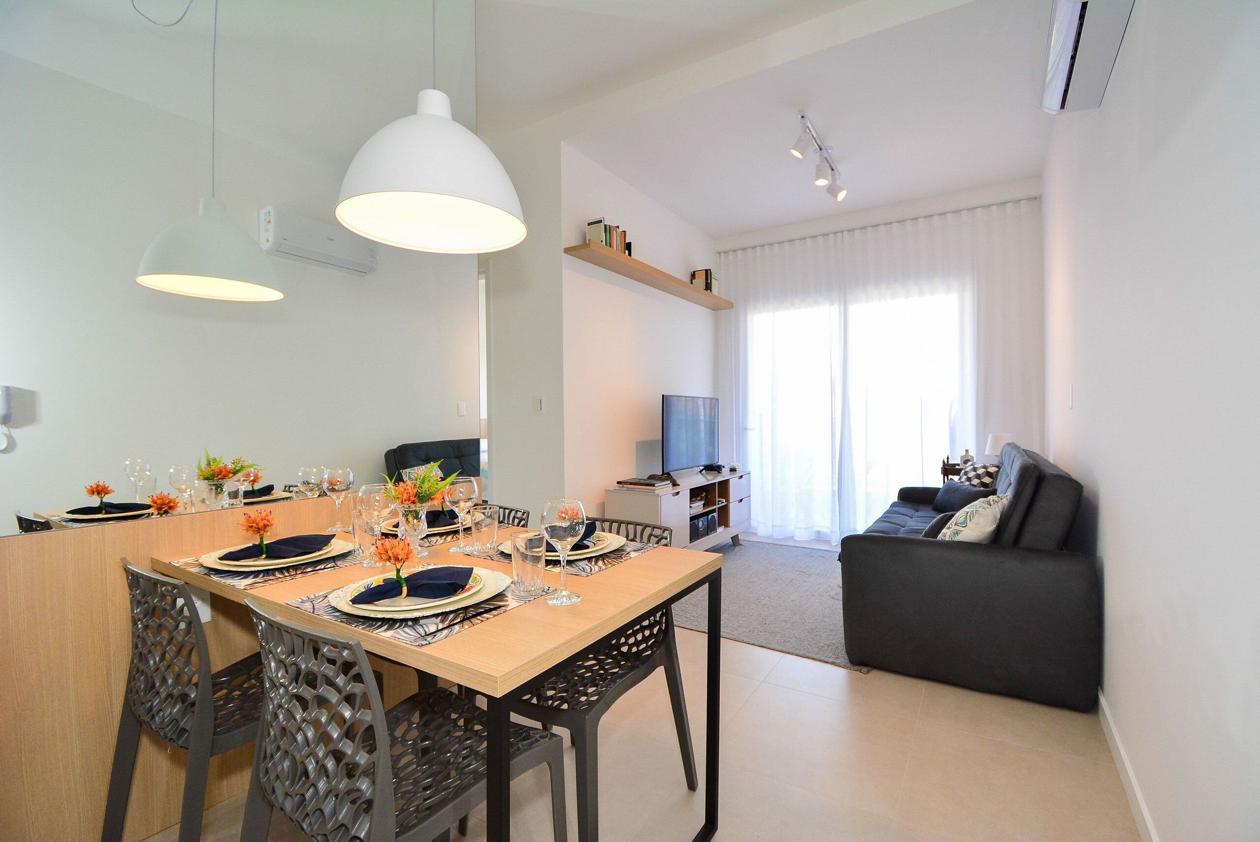 A modern dining area with a wooden table set for four, surrounded by chairs. A television is mounted on the wall, and a sofa is visible in the background.