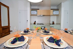 A dining table set for four with floral centerpieces, a microwave above the counter, and a wooden door to the left. - SunClub-300 metros da Praia do Campeche (Florianopolis)