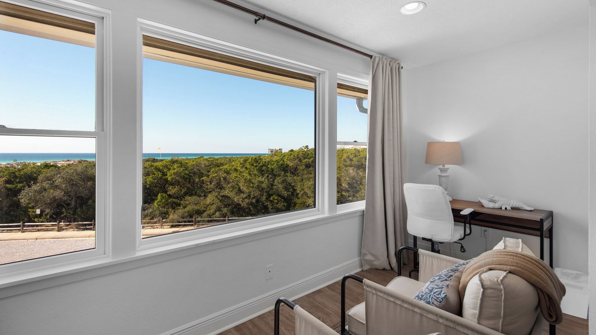 Una habitación con un amplio ventanal que ofrece vistas a la playa y al océano. Hay un escritorio con una lámpara, una silla y una cama con una manta.