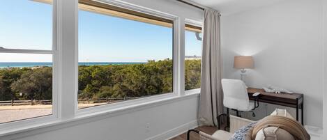 Una habitación con un amplio ventanal que ofrece vistas a la playa y al océano. Hay un escritorio con una lámpara, una silla y una cama con una manta.