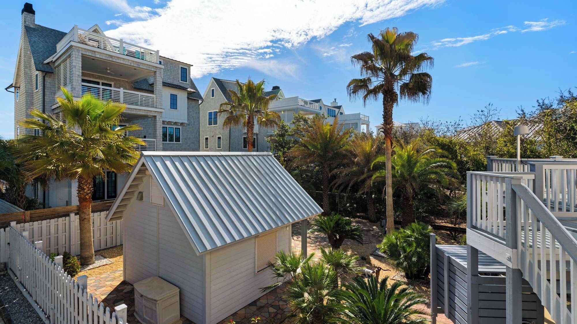A residential area with multiple houses, including a small shed with a corrugated metal roof, surrounded by palm trees and a white picket fence.