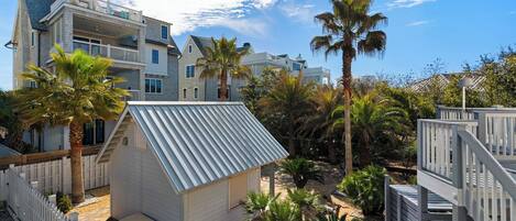 A residential area with multiple houses, including a small shed with a corrugated metal roof, surrounded by palm trees and a white picket fence.