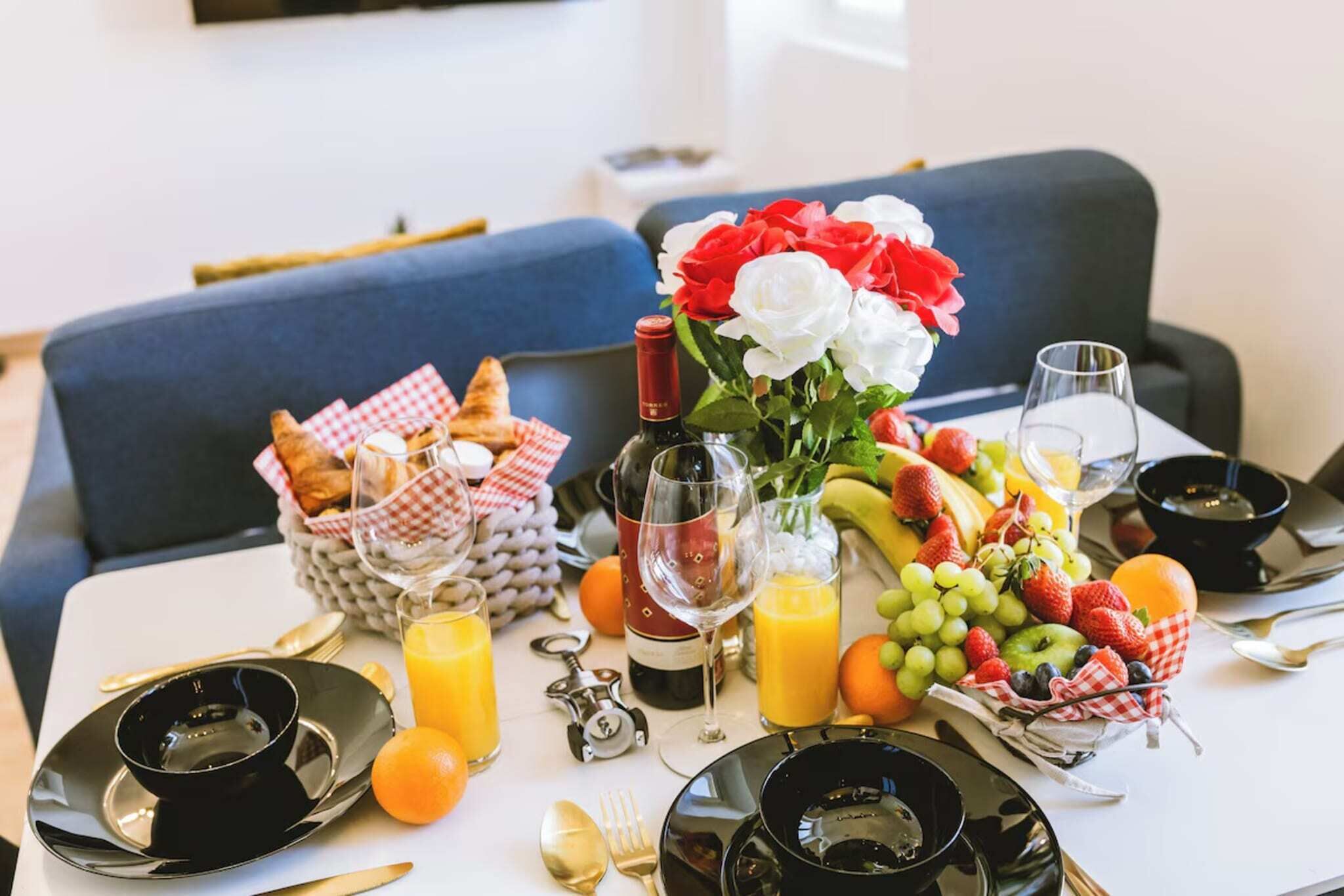 A breakfast table with croissants, a bottle of wine, a vase of flowers, and a fruit platter.