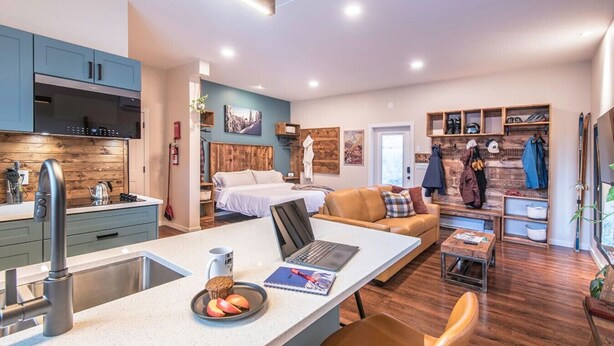 A modern kitchen with a central island, a dining area with a table and chairs, a bedroom with a bed and nightstands, and a living area with a sofa and a coffee table. - Sparkling NEW Rossland Midtown Studio Apartment (Rossland)