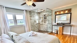 A hotel room with a bed, a shower with glass doors, a vanity with a mirror, and a window with curtains.