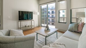 A modern living room with a flat-screen TV, a sofa, a coffee table, and a balcony with a view of a building. - Landing Apartments Houston (Houston)