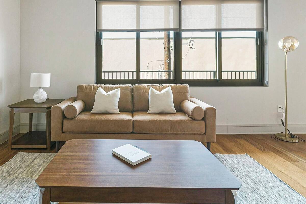Een woonkamer met een bruine leren bank, een houten bijzettafeltje, een nachtkastje met een lamp en een vloerlamp.