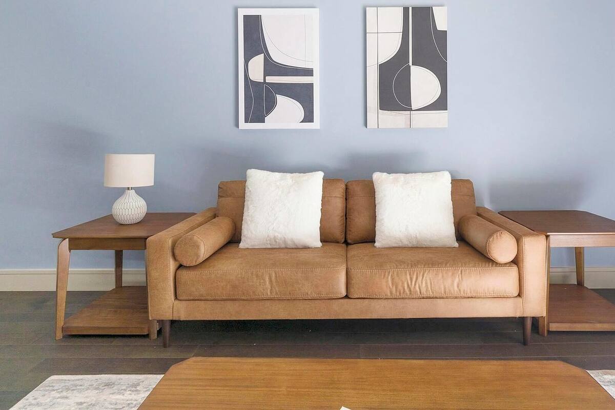 Een bruine leren bank met twee witte kussens, een kleine bijzettafel met een lamp, en twee abstracte schilderijen aan de muur.