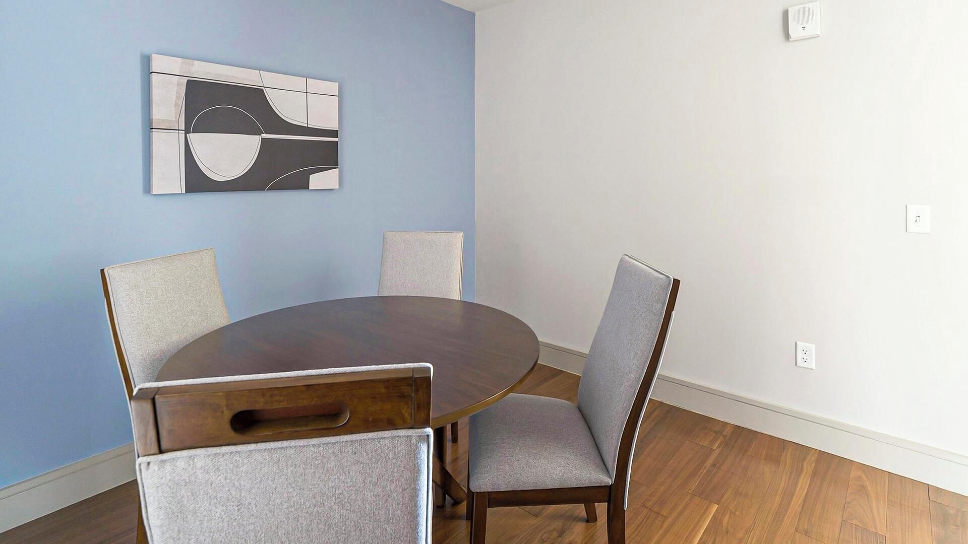 A dining area with a round table, four chairs, a wall-mounted abstract art piece, and a smoke detector.