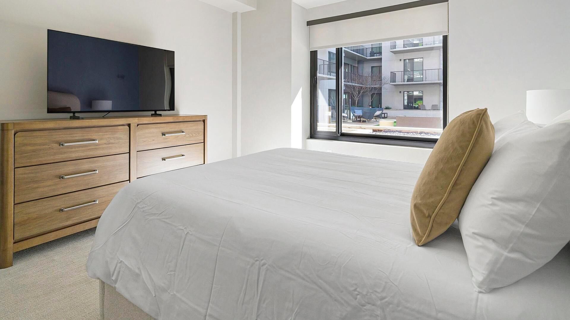 A bedroom with a bed, a TV on a wooden dresser, and a window with a view of buildings.