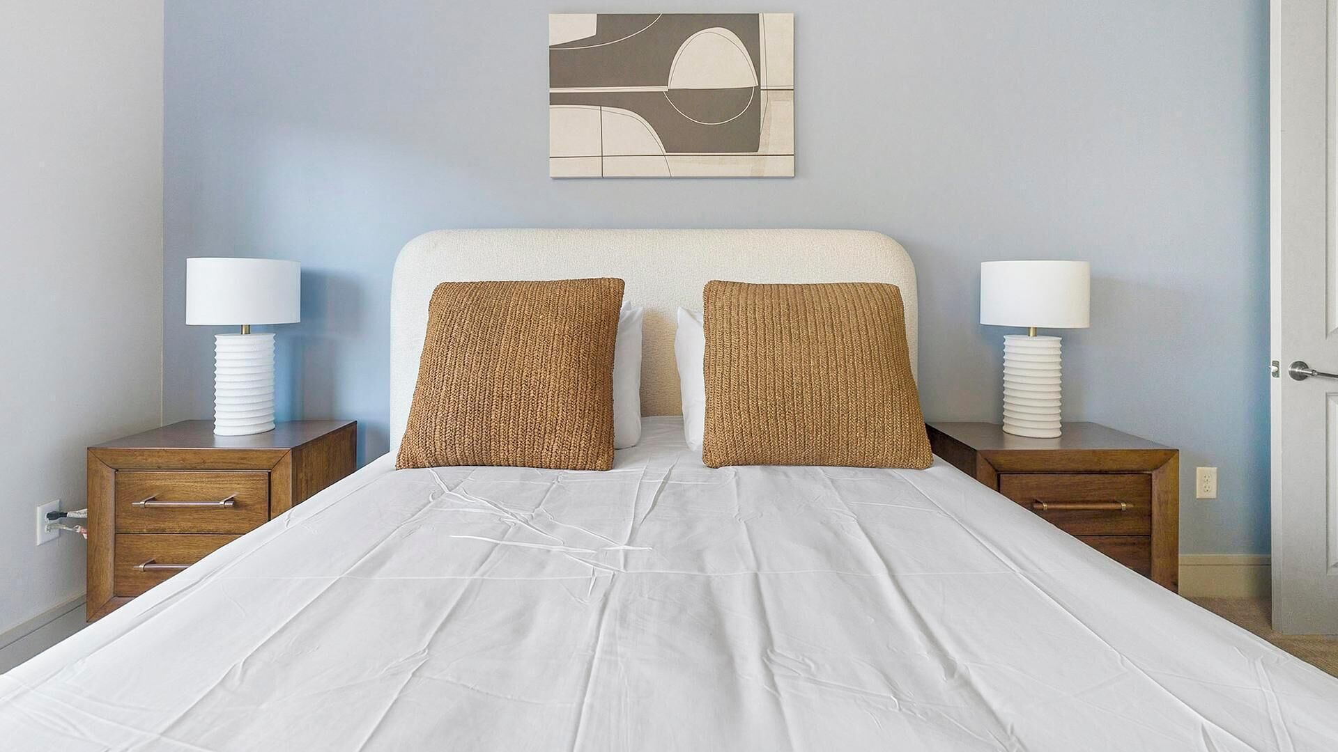 A neatly made bed with white linens and two textured pillows, flanked by wooden nightstands with white lamps.