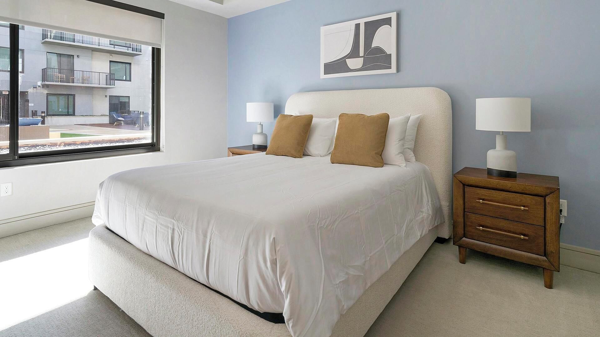 A bedroom with a large bed, two bedside tables, a window with a view of a building, and a wall-mounted artwork.