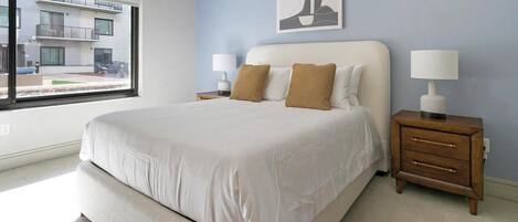 A bedroom with a large bed, two bedside tables, a window with a view of a building, and a wall-mounted artwork.