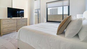 A hotel room with a bed, a TV on a wooden dresser, and a large window with a city view. - Landing Apartments Houston (Houston)