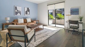 A modern living room with a sofa, coffee table, and a desk with a chair. There are two abstract paintings on the wall, a large window with a view of greenery, and a floor lamp. - Landing Apartments Houston (Houston)