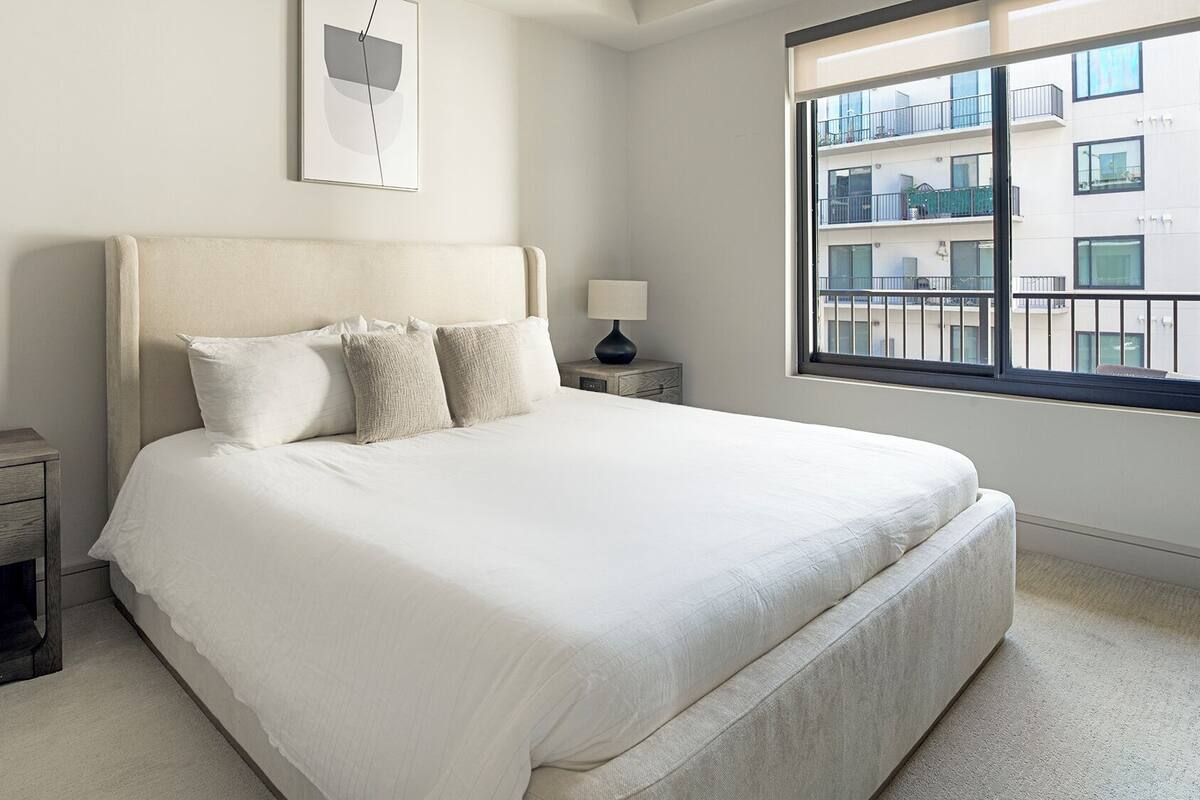 Een slaapkamer met een groot bed, twee nachtkastjes en een raam met uitzicht op gebouwen.