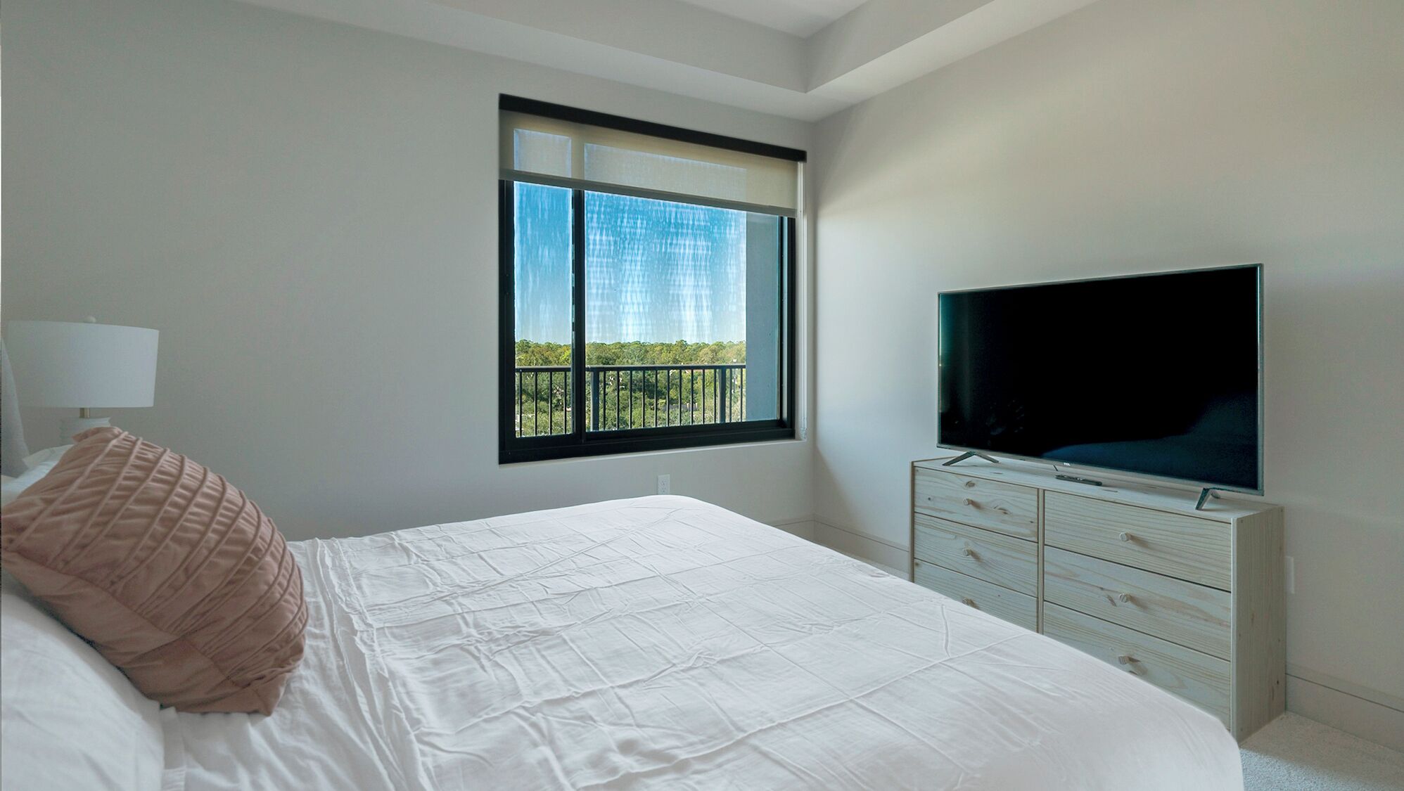 A bedroom with a large bed, a TV on a dresser, a window with a view of greenery, and a lamp on a bedside table.
