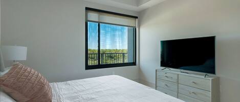 A bedroom with a large bed, a TV on a dresser, a window with a view of greenery, and a lamp on a bedside table.