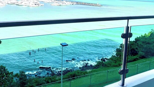 Vista costanera des del balcó, amb cel blau, un port esportiu amb embarcacions i un turó allunyat amb edificis.