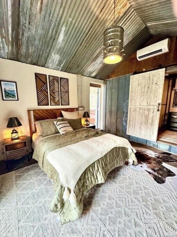 A rustic bedroom with a wooden bed, a nightstand with a lamp, a wall-mounted air conditioner, and a door with a rustic design.