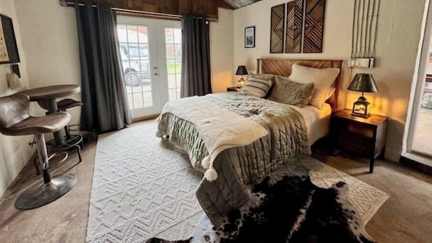 A bedroom with a bed, a desk, and a chair. There is a window with curtains, a wall-mounted lamp, and a rug on the floor.