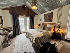 A bedroom with a bed, a desk, and a chair. There is a window with curtains, a wall-mounted lamp, and a rug on the floor.