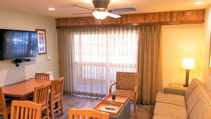 A hotel room with a dining table, chairs, a sofa, a TV, a ceiling fan, and a large window with curtains.