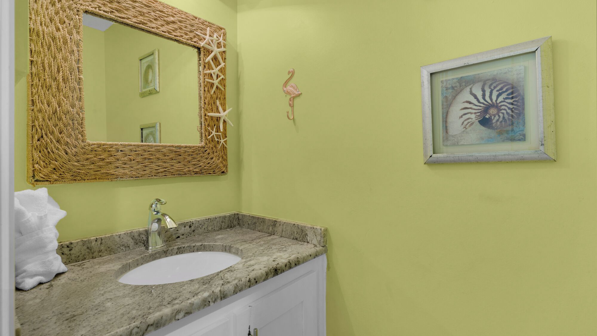 A bathroom with a granite countertop, a white sink, a woven mirror frame, and a framed seahorse painting on the wall.