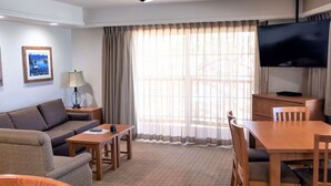 Ein Wohnzimmer mit einer Couch, einem Tisch, einem Fernseher und einem großen Fenster mit Vorhängen.