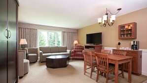 A hotel room with a living area, a dining table with chairs, a sofa, a television, a coffee maker, and a cabinet with glassware. - Deluxe Studio | Murphy Bed + Sofa (West Dover)